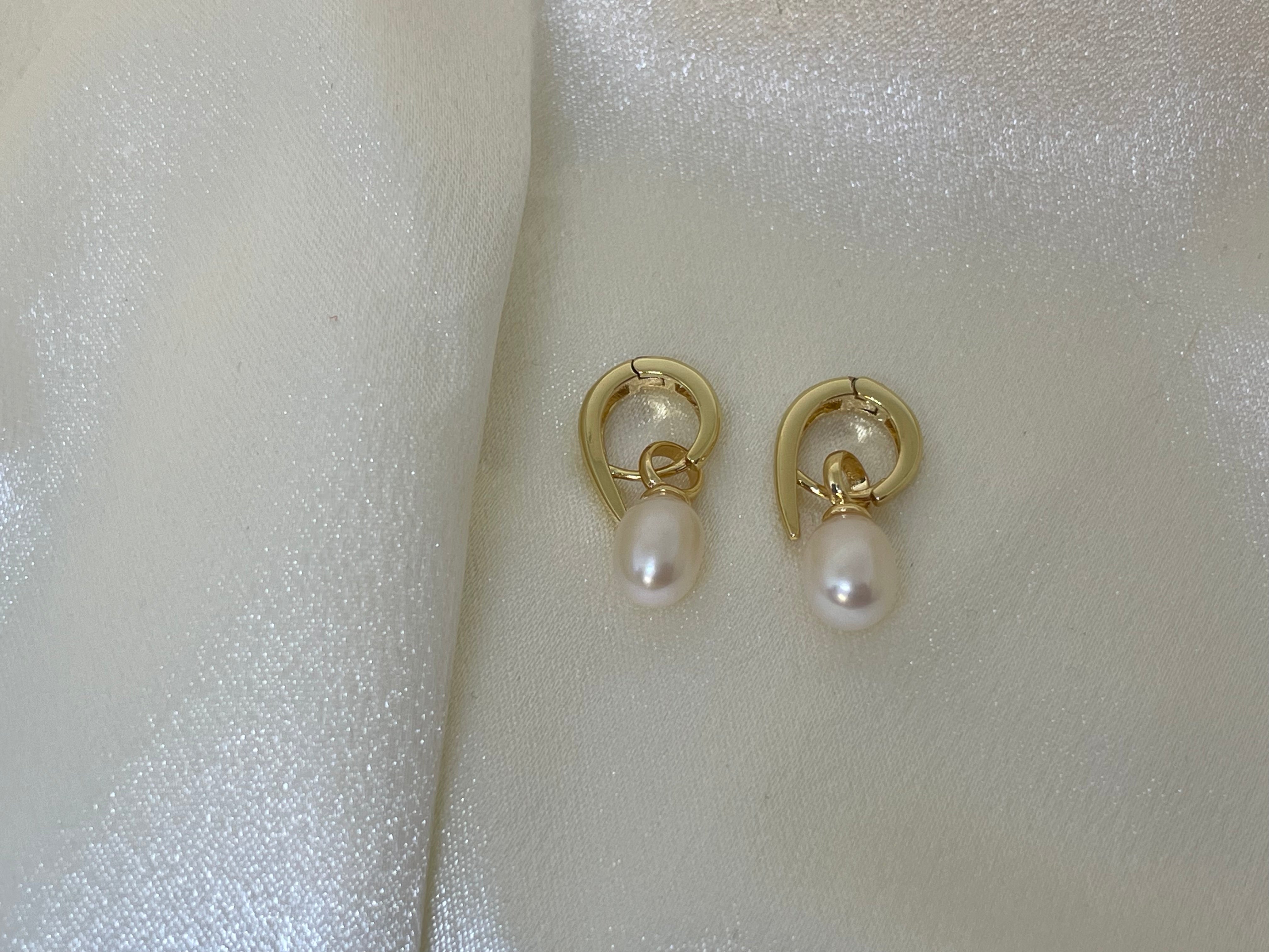 Two gold twist earrings with freshwater pearl drops are displayed on a white, shimmery fabric.
