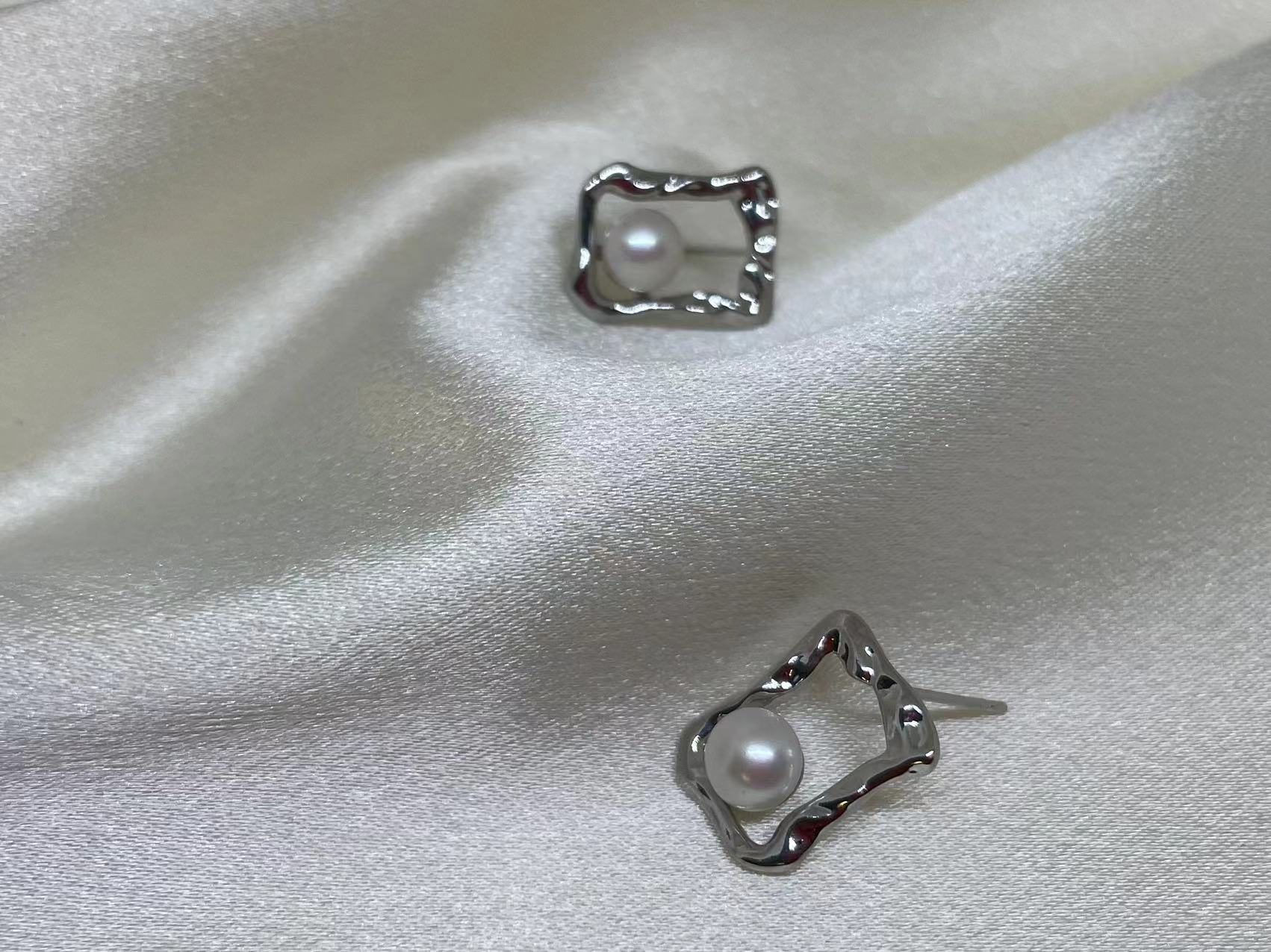 Two silver quadrate stud earrings with freshwater pearls are displayed on white silk.