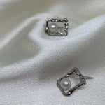 Two silver quadrate stud earrings with freshwater pearls are displayed on white silk.