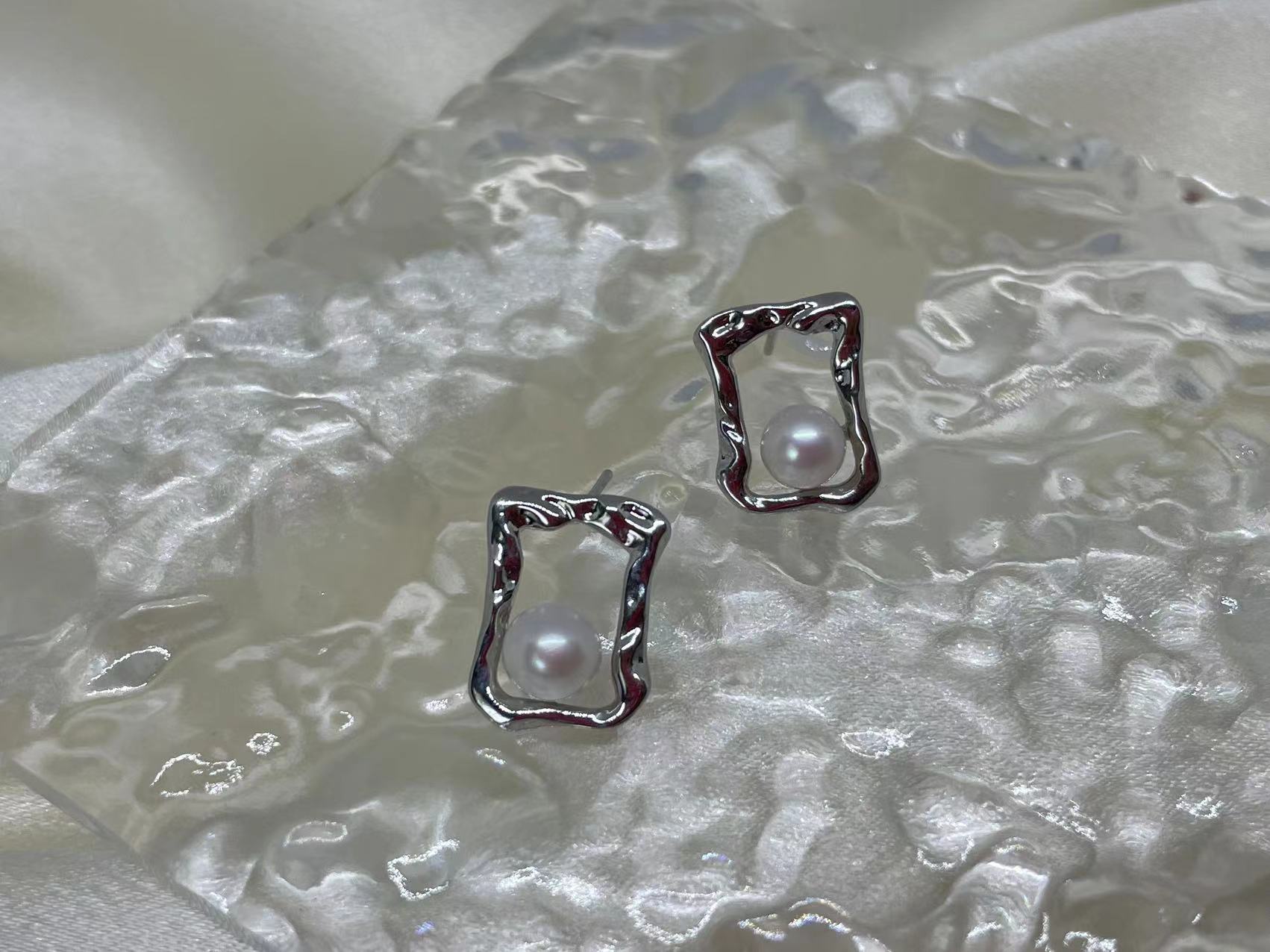 Two silver quadrate stud earrings with freshwater pearls are displayed on a textured surface.