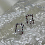 Two silver quadrate stud earrings with freshwater pearls are displayed on a textured surface.