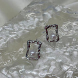 Two silver quadrate stud earrings with freshwater pearls are displayed on a textured surface.
