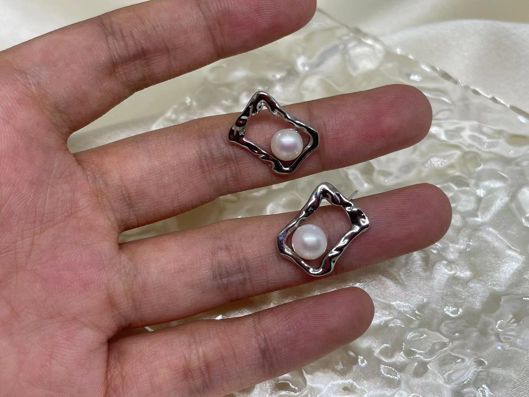 Two silver quadrate stud earrings with freshwater pearls are held in a hand.