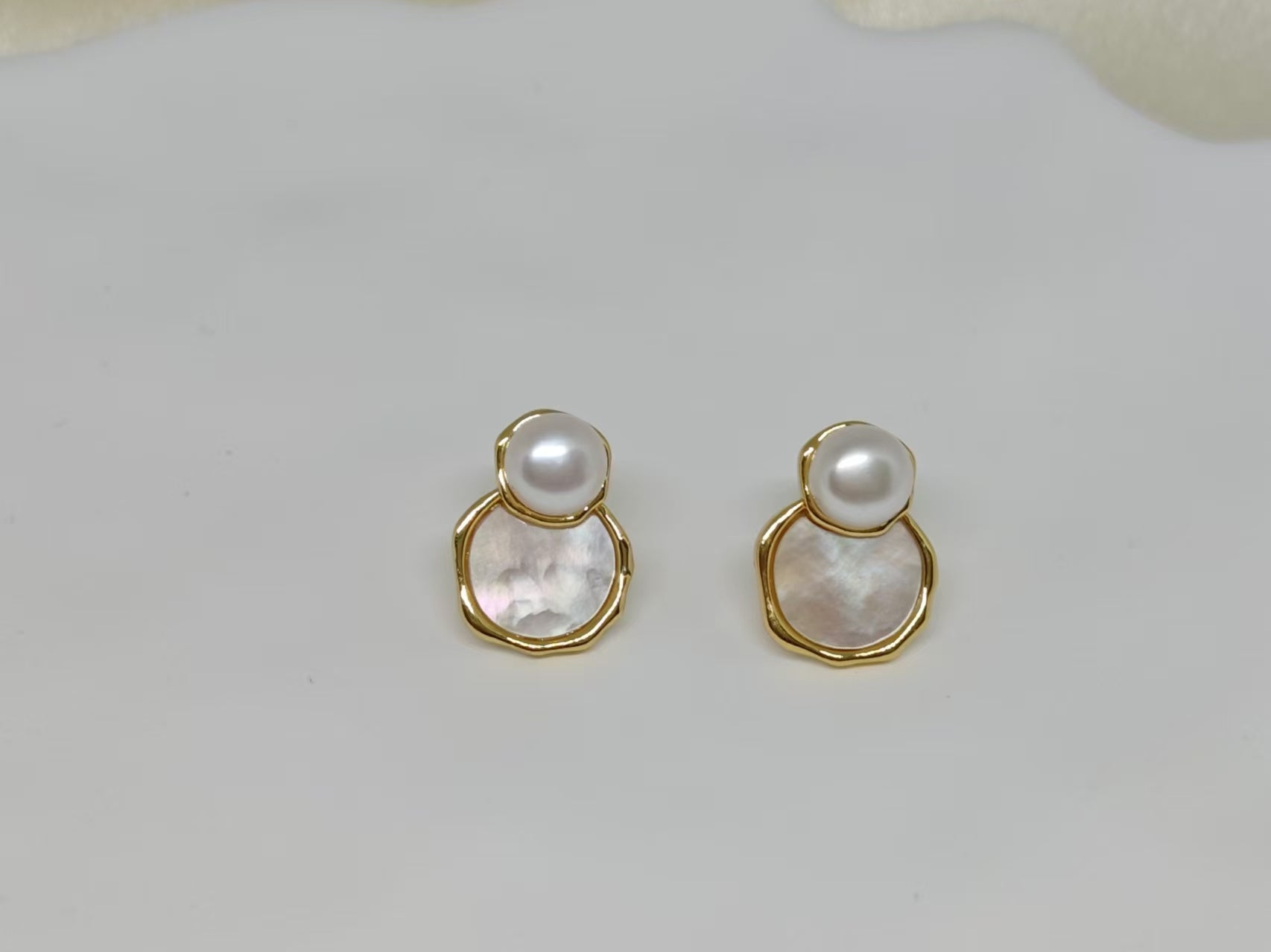 Two gold-framed earrings with a white pearl atop a mother-of-pearl disc.