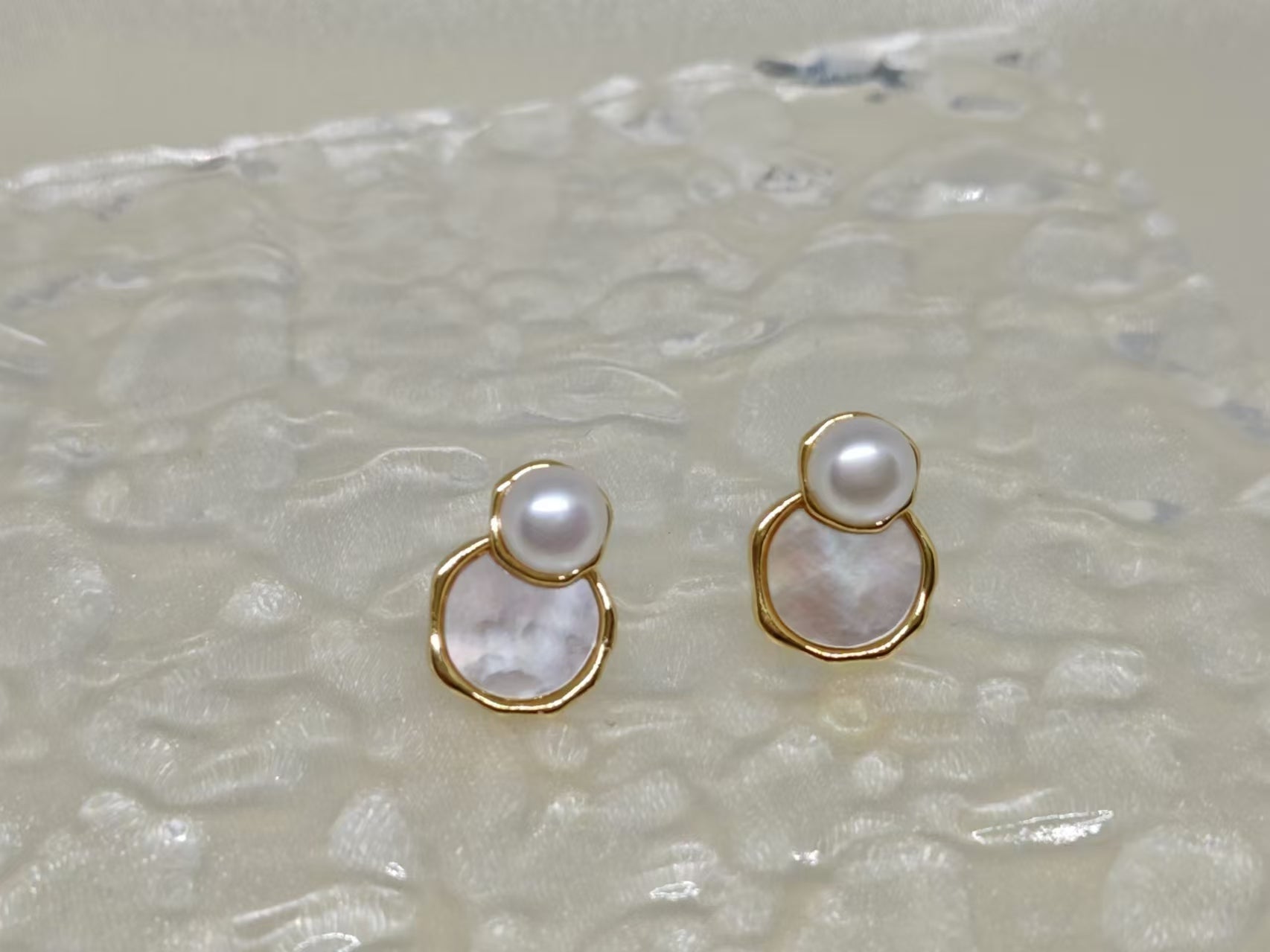 Two gold-rimmed earrings, each with a white pearl atop a mother-of-pearl disc.