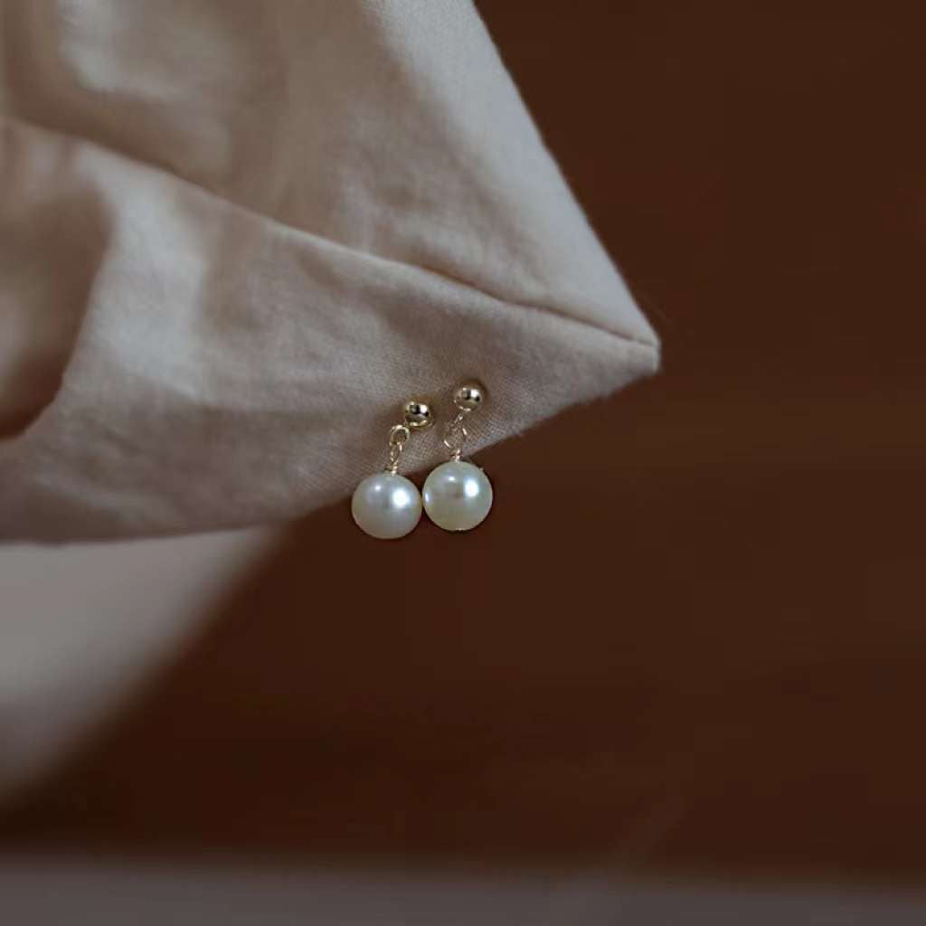 Small round pearl dangle earrings with gold-plated studs are displayed on a light fabric.