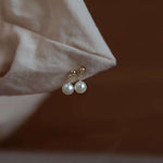 Small round pearl dangle earrings with gold-plated studs are displayed on a light fabric.