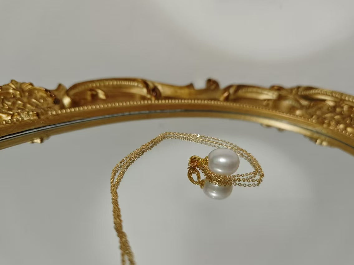A gold chain necklace with a large, off-round white pearl pendant, reflected on a mirror.