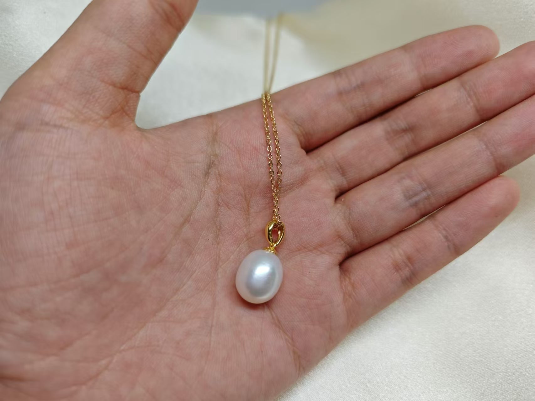 A white freshwater pearl pendant on a delicate gold chain held in a hand.