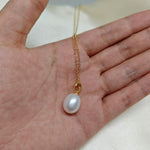A white freshwater pearl pendant on a delicate gold chain held in a hand.
