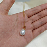A white freshwater pearl pendant on a delicate gold chain held in a hand.