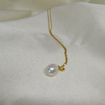 A white, off-round freshwater pearl hangs from a delicate gold chain on a white fabric.