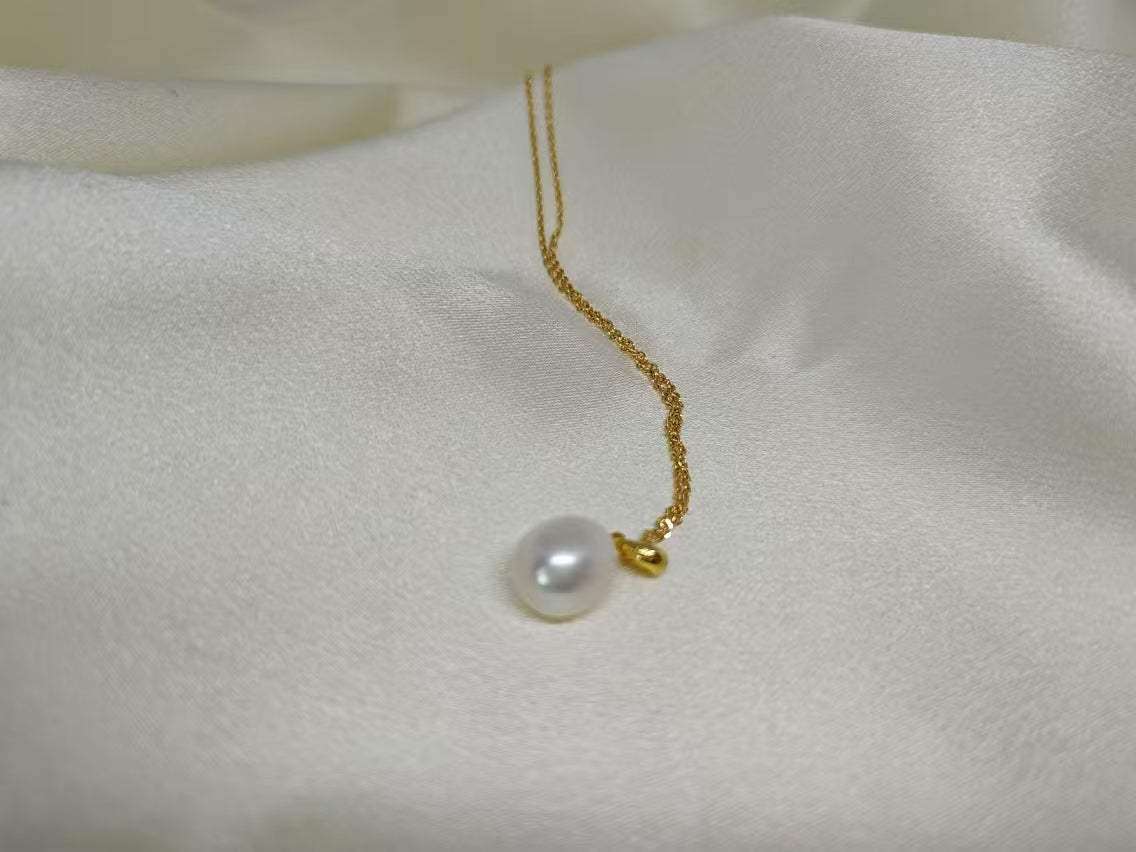 A white, off-round freshwater pearl hangs from a delicate gold chain on a white fabric.