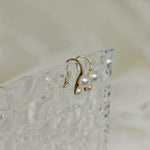 Gold-plated freshwater pearl stud earrings with curved hooks on a textured clear surface.