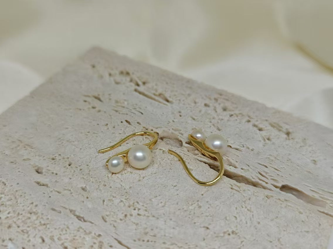 Gold-plated freshwater pearl stud earrings with curved hooks on a textured surface.