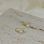 Gold-plated freshwater pearl stud earrings with curved hooks on a textured surface.