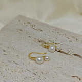 Gold-plated freshwater pearl stud earrings with curved hooks on a textured surface.
