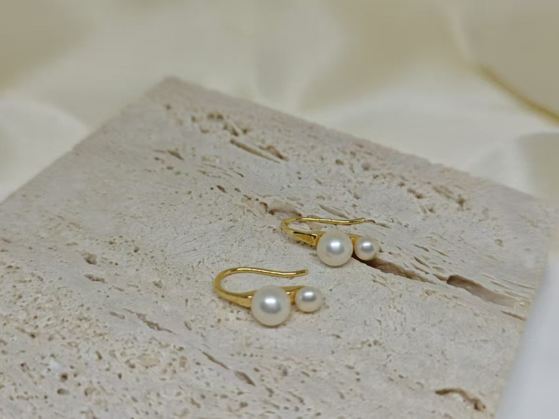 Gold-plated freshwater pearl stud earrings with curved hooks on a textured surface.