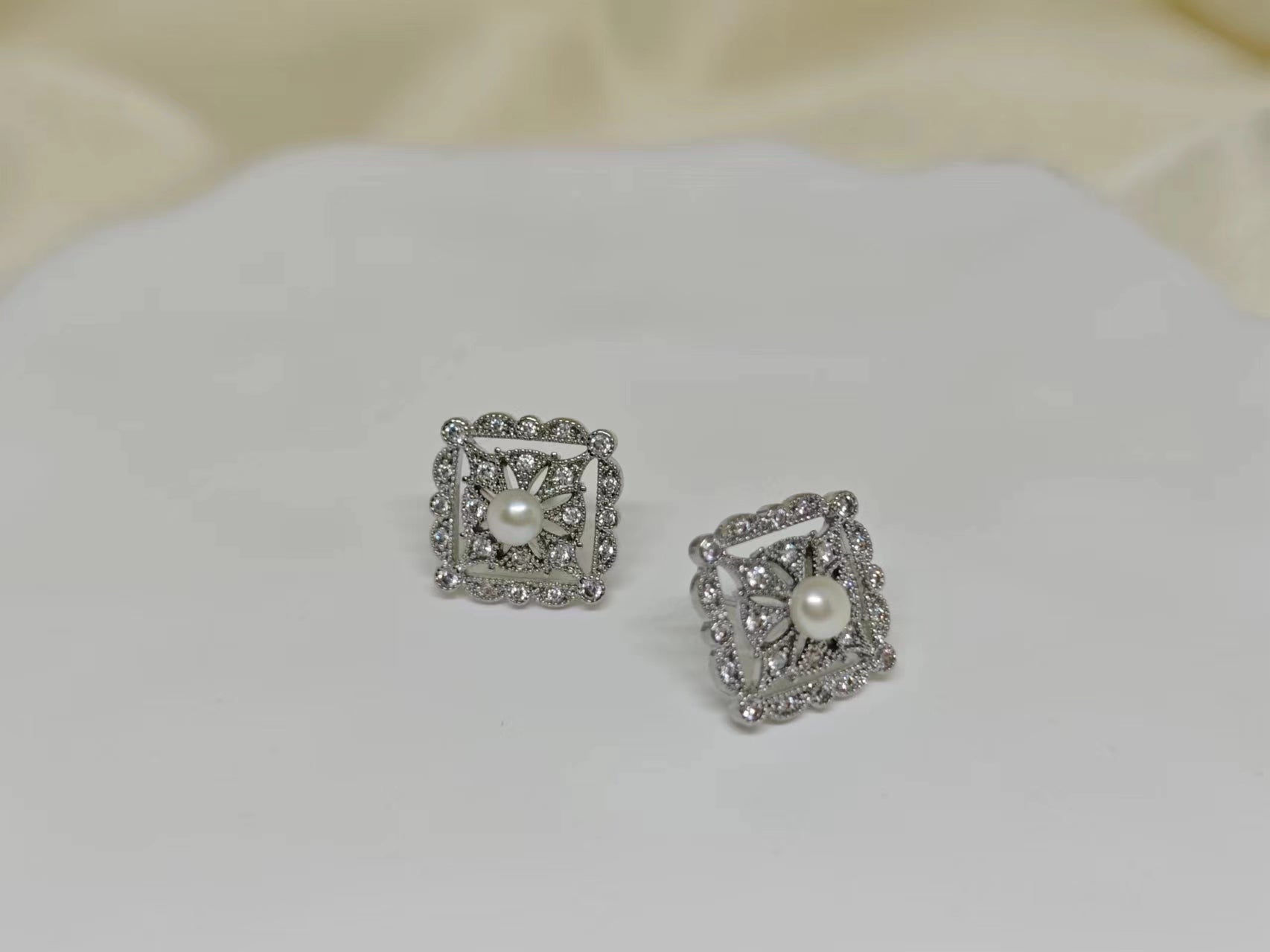 A pair of silver square stud earrings with a central pearl surrounded by small diamonds.