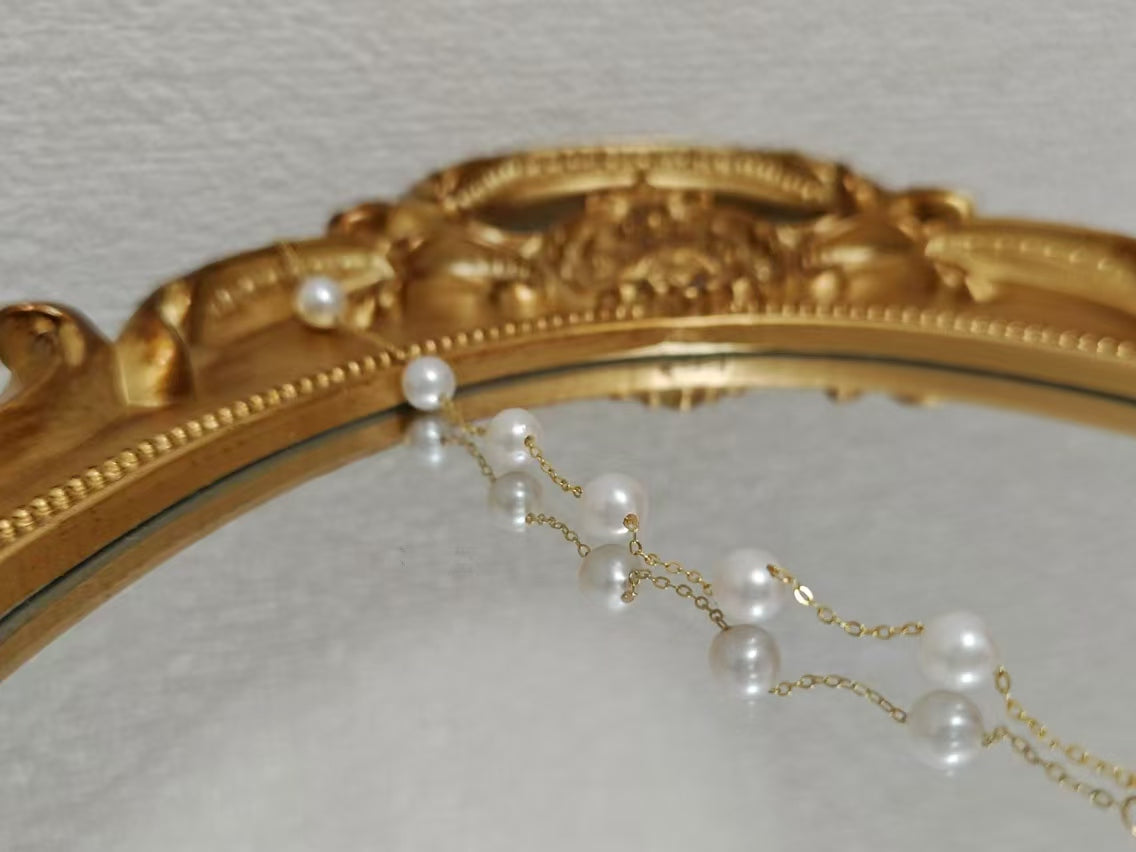Seven freshwater pearls are strung on a delicate gold chain, reflected in a mirror.