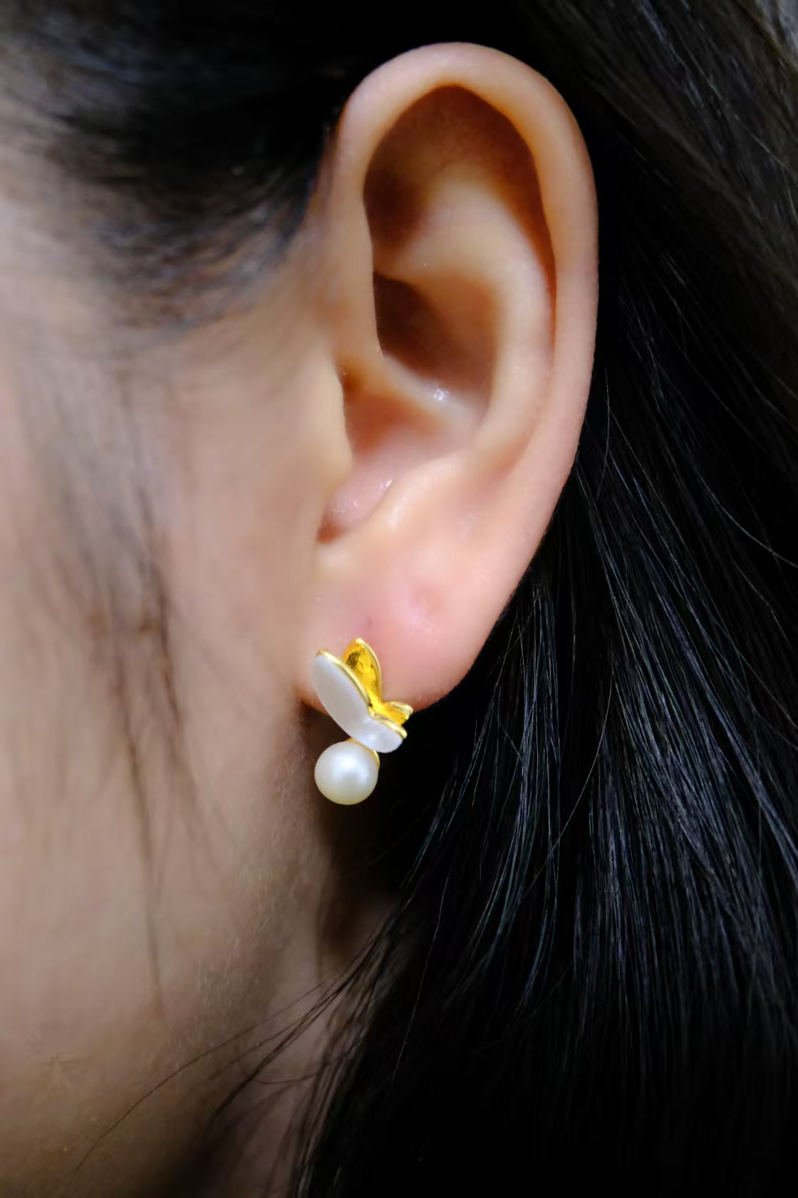A gold-plated butterfly stud earring with a freshwater pearl dangles from an ear.