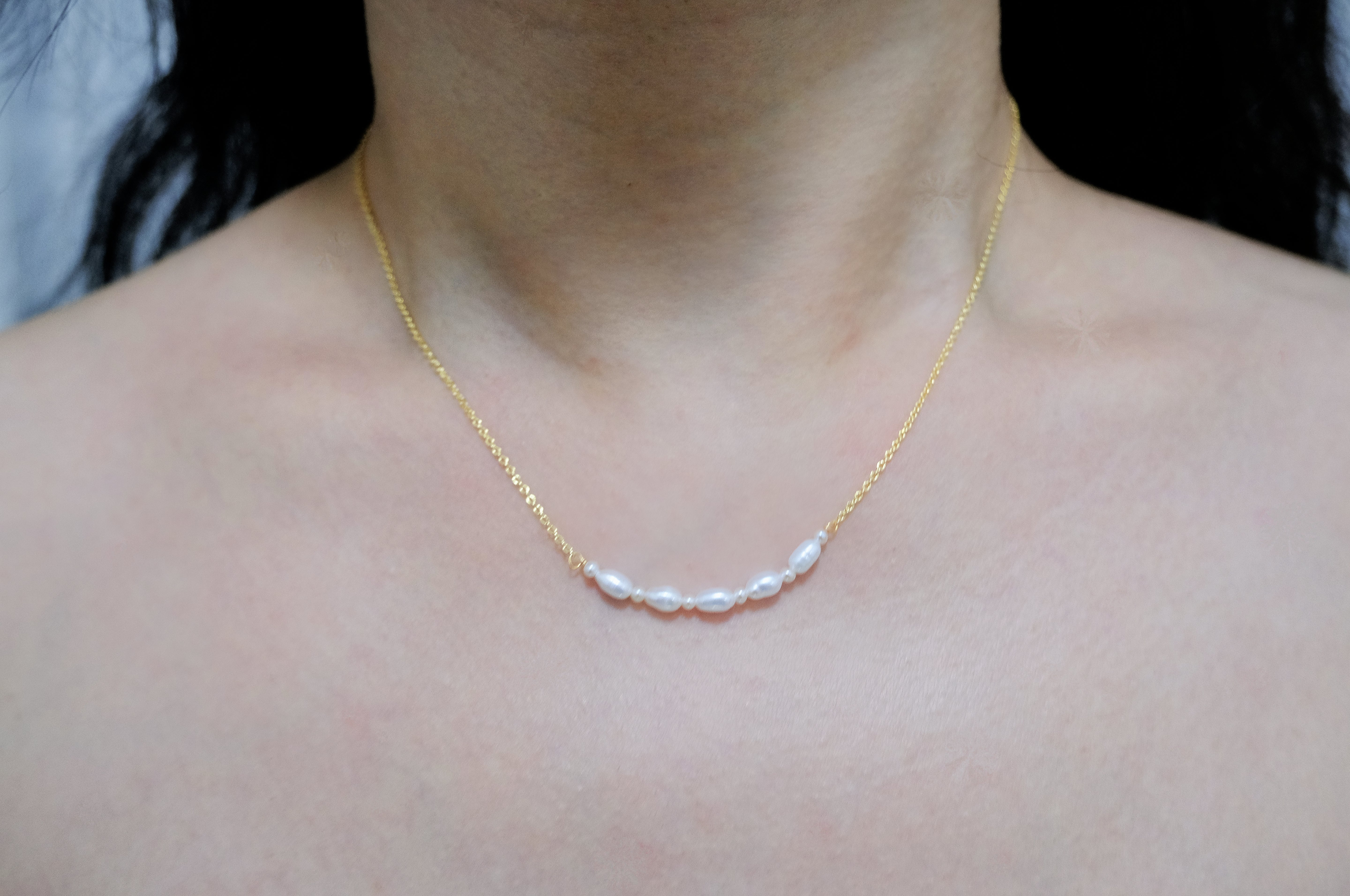 A woman wears a gold chain necklace with five freshwater pearls across her collarbone.