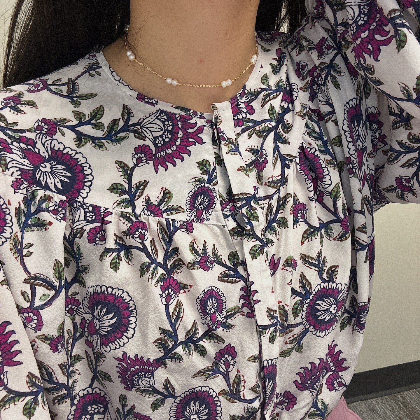 A woman wears a gold-plated pearl necklace with a white floral blouse.