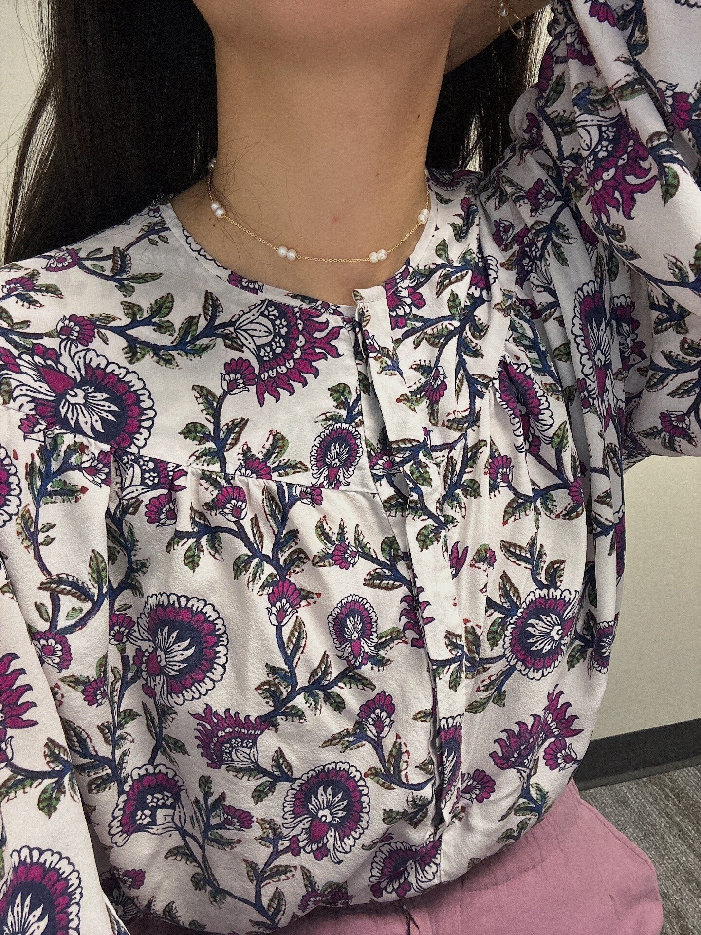 A woman wears a gold-plated pearl necklace with a white floral blouse.