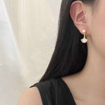 A person wears Caellia 14K gold earrings featuring a freshwater pearl stud and a mother-of-pearl fan.