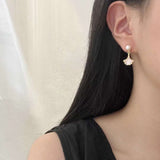 A person wears Caellia 14K gold earrings featuring a freshwater pearl stud and a mother-of-pearl fan.