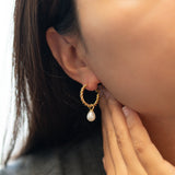 A person wears a Caellia 14K gold twisted hoop earring with a dangling freshwater pearl.