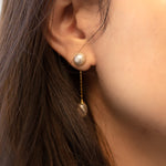 A close-up of a person wearing CAELLIA gold-plated pearl dangle earrings with a teardrop design.