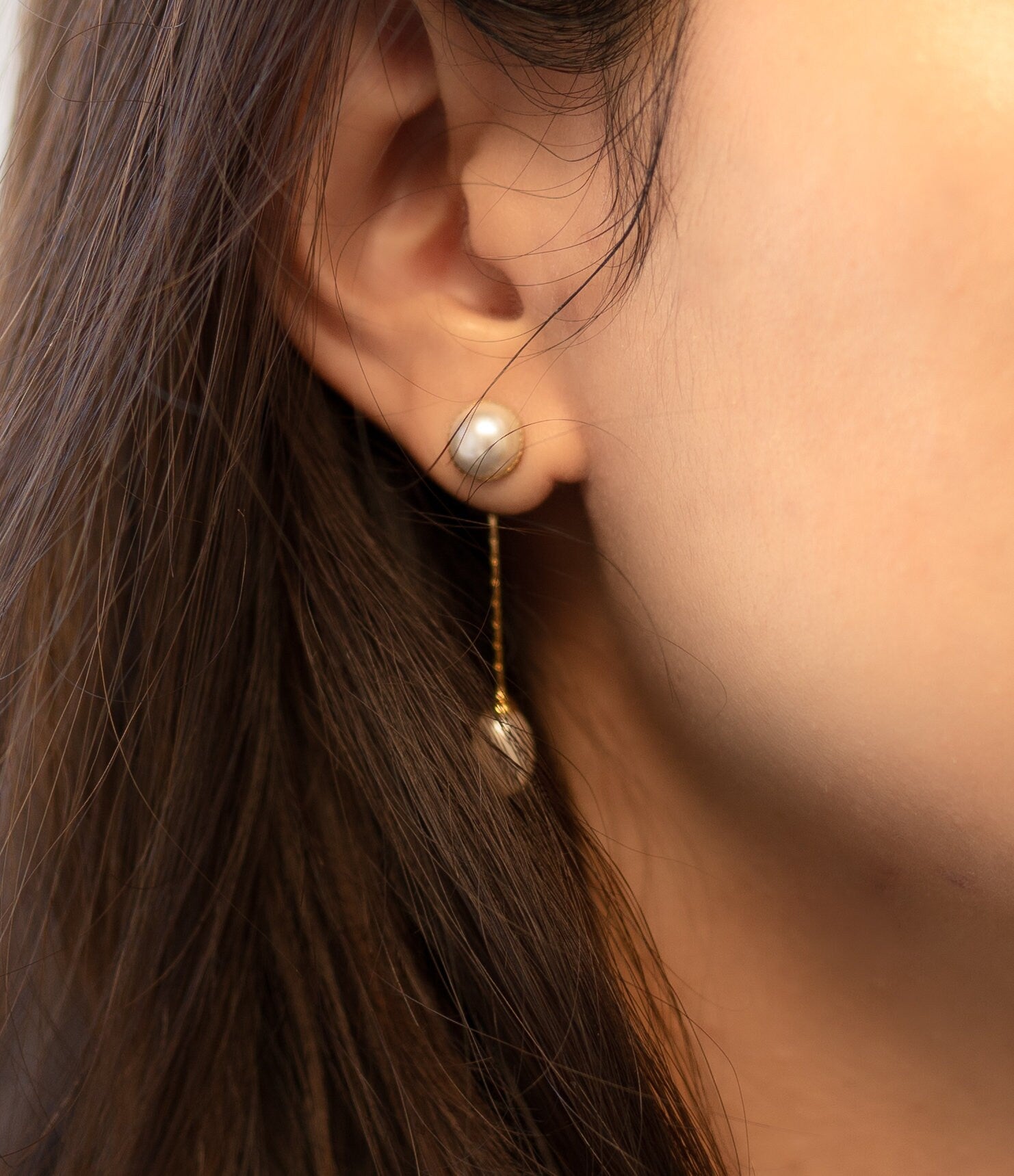 A close-up of a person wearing CAELLIA gold-plated pearl dangle earrings with a teardrop design.
