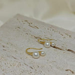 Caellia 18K gold plated stud earrings featuring two freshwater pearls on a textured stone surface.