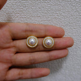 A pair of CAELLIA gold plated freshwater pearl earrings with a circular frame held in a hand.