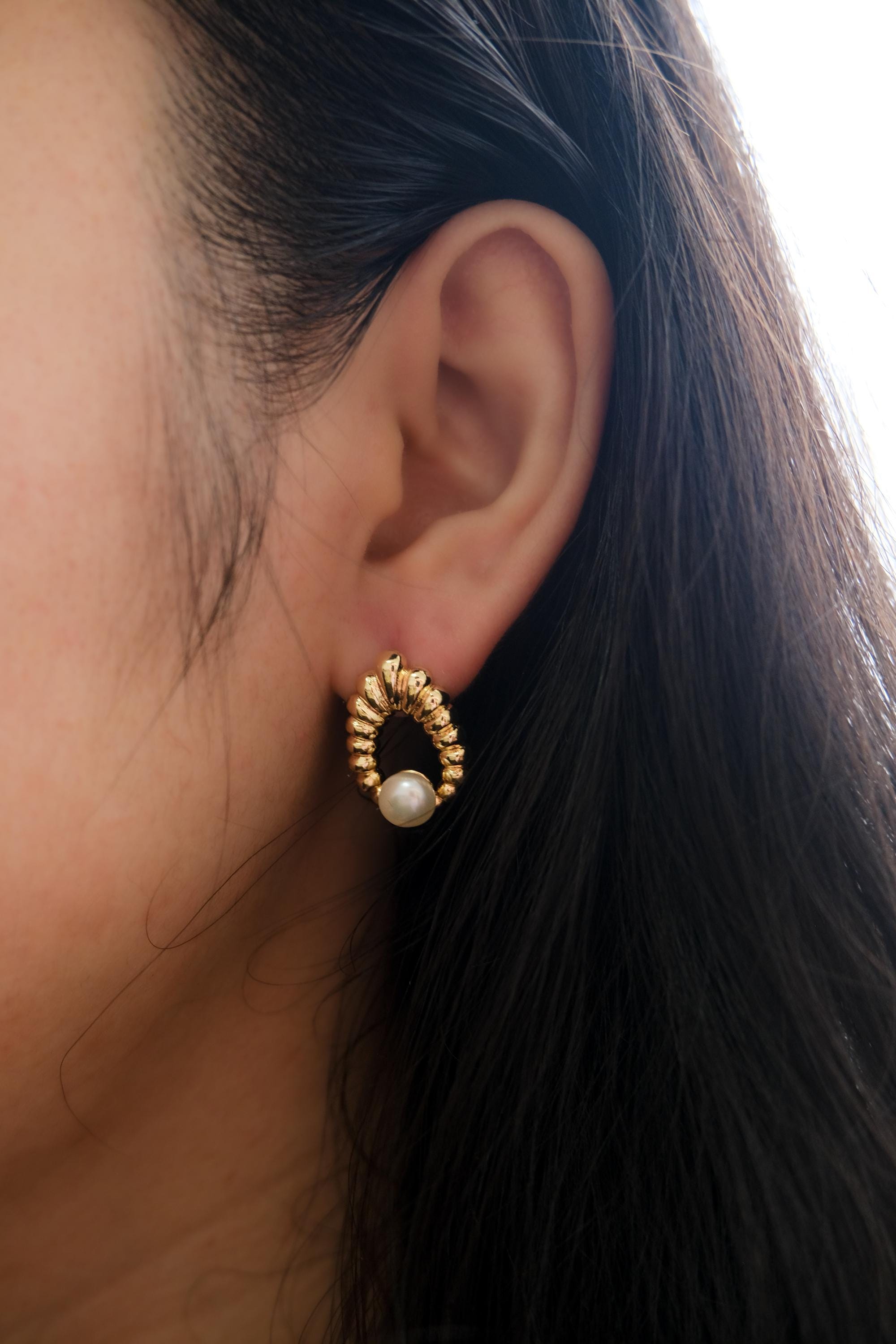 A person wears a vintage-style CAELLIA gold earring featuring a textured hoop and a pearl drop.
