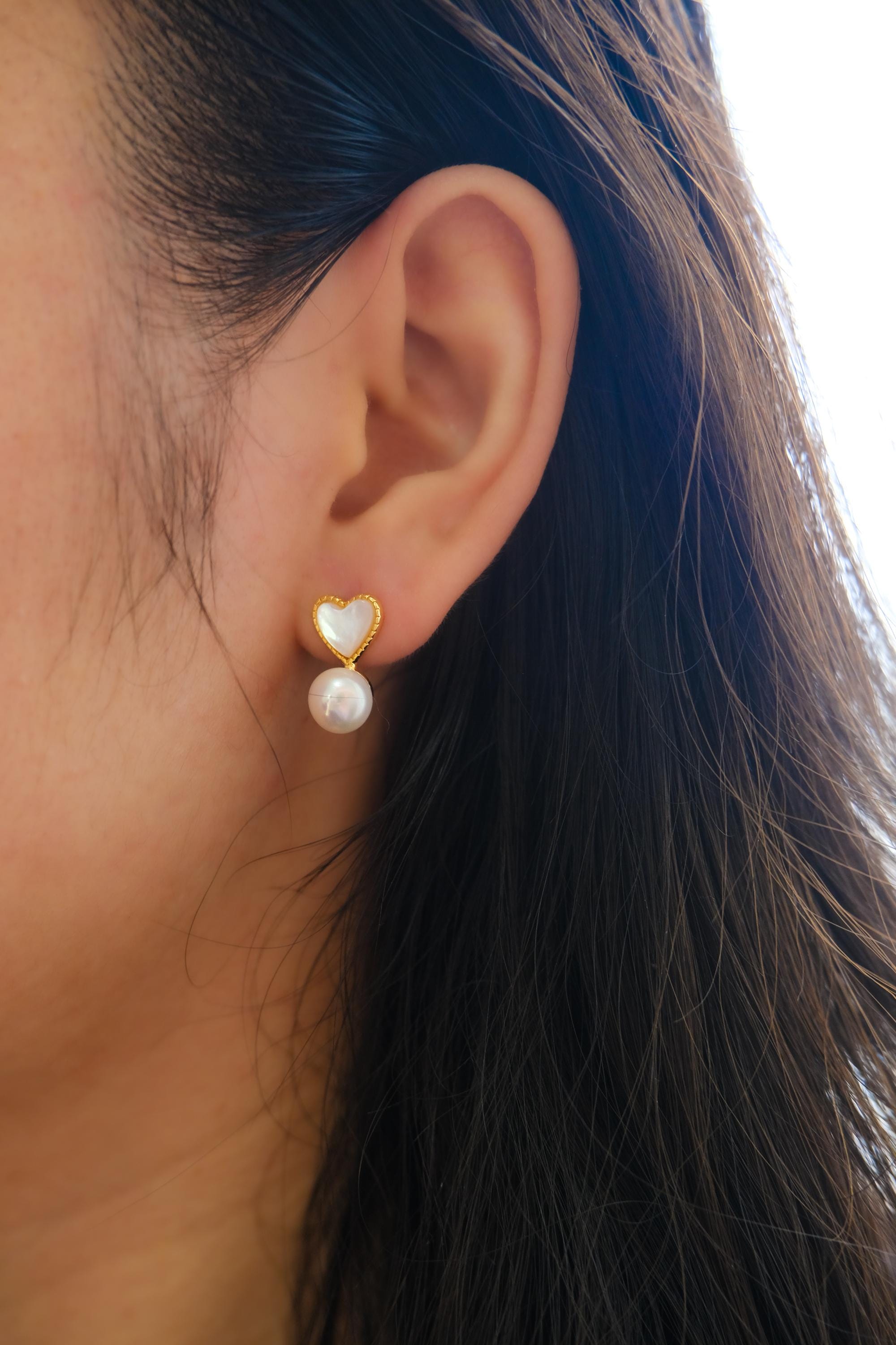 A person wears Caellia 14K gold heart-shaped fritillaria shell and freshwater pearl stud earrings.