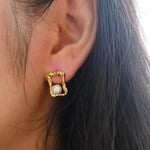 A person wears a CAELLIA gold plated stud earring featuring a freshwater pearl in a quadrate frame.