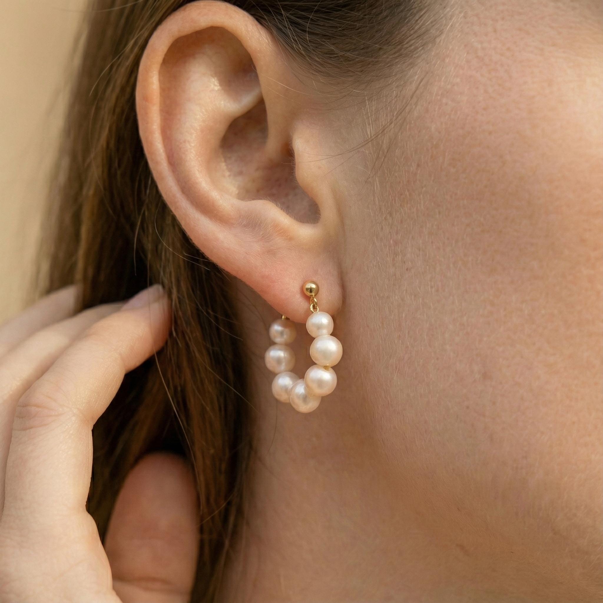 A person wears Caellia 18K gold hoop earrings featuring a row of small freshwater pearls.
