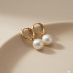 Caellia 18K gold hoop earrings featuring luminous freshwater pearls resting on a beige surface.