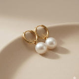 Caellia 18K gold hoop earrings featuring luminous freshwater pearls resting on a beige surface.