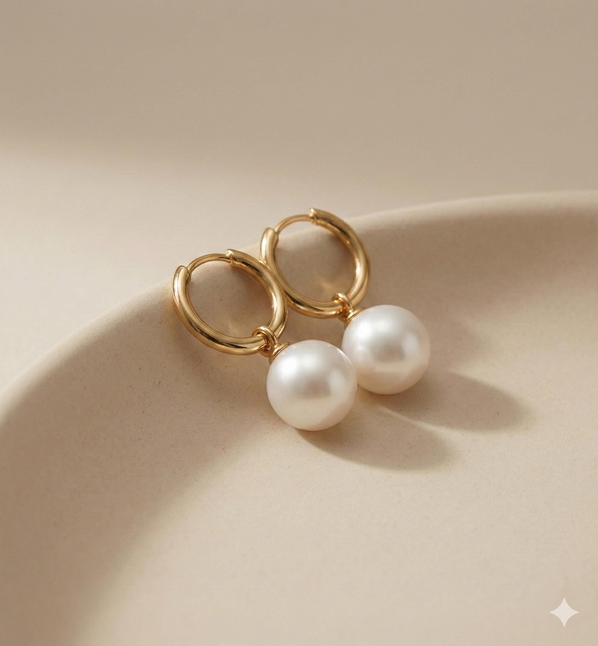 Caellia 18K gold hoop earrings featuring luminous freshwater pearls resting on a beige surface.