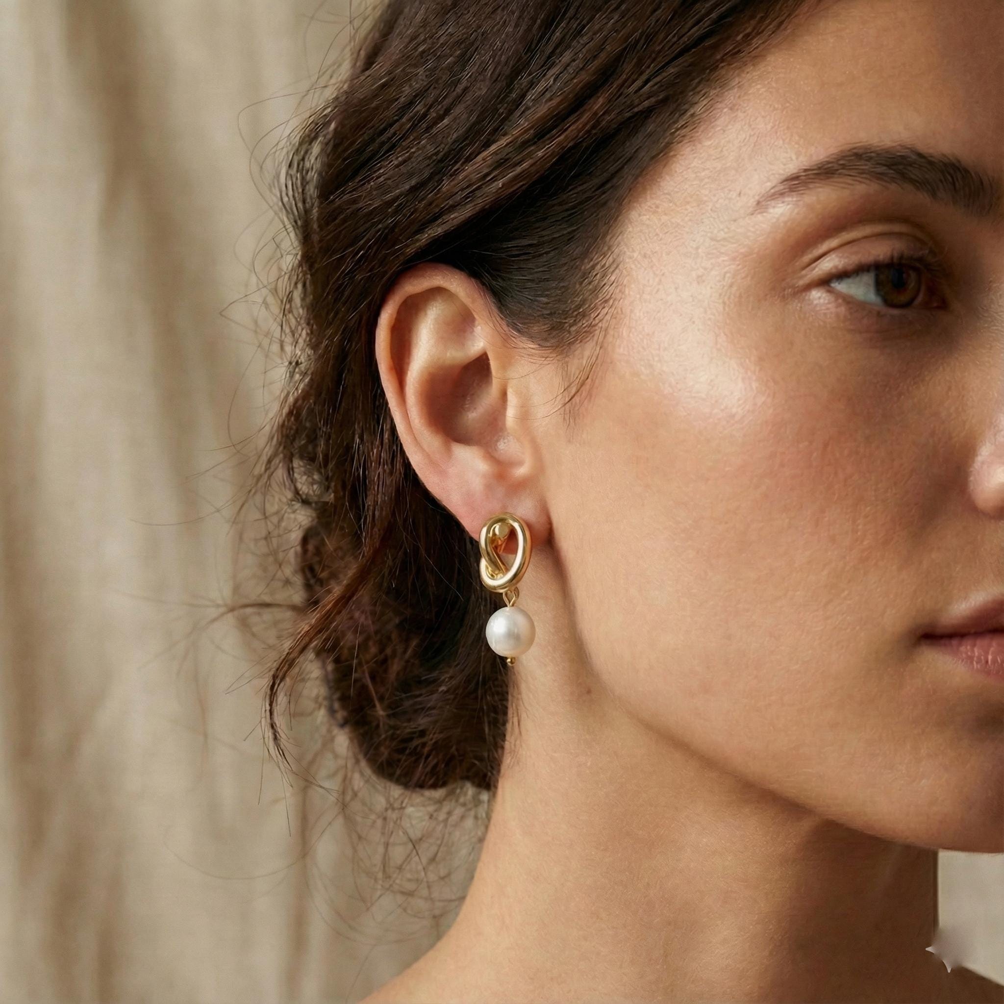 A woman wears Caellia 14K gold knot earrings featuring a dangling freshwater pearl.