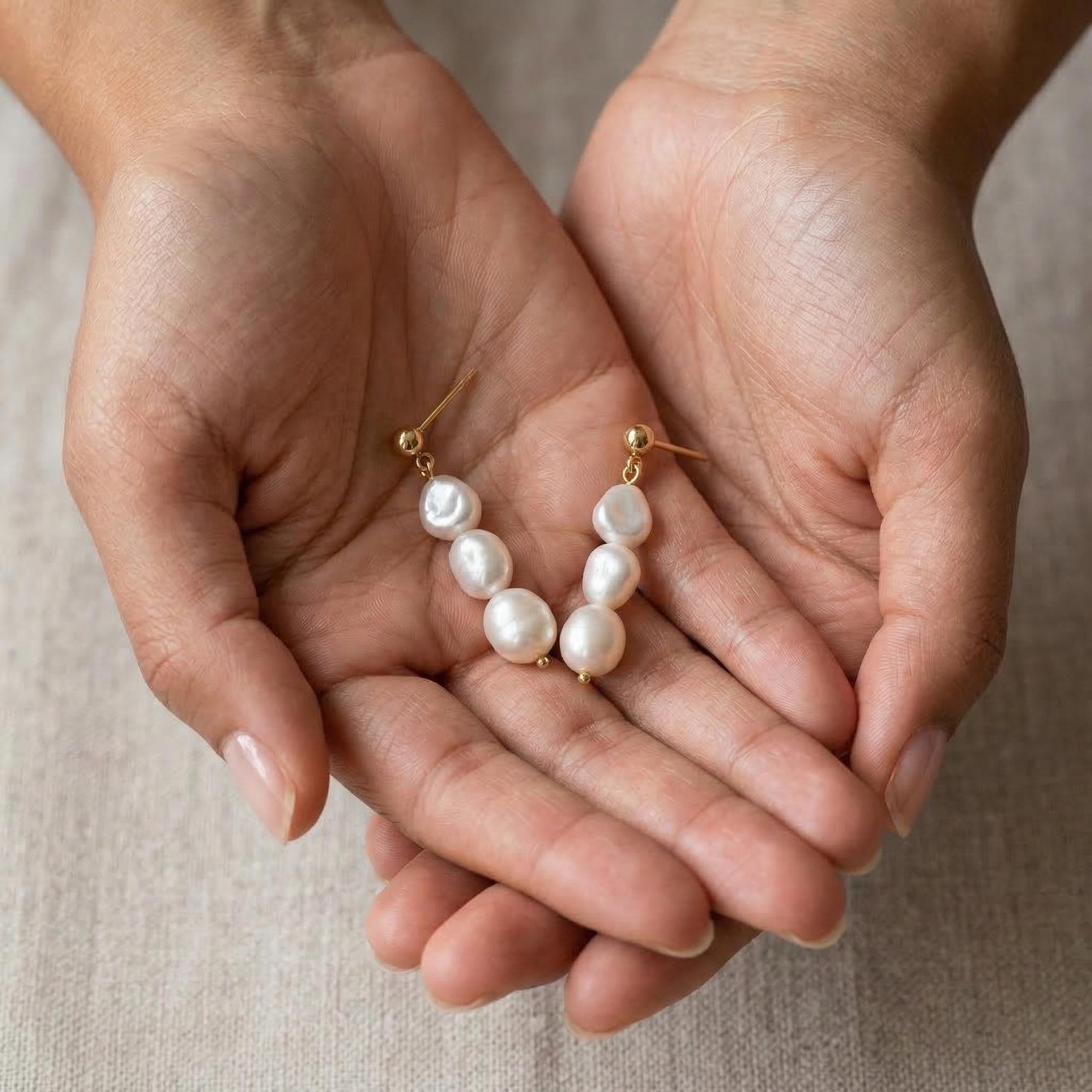 Caellia 14K gold triple freshwater pearl drop earrings held in a pair of hands.