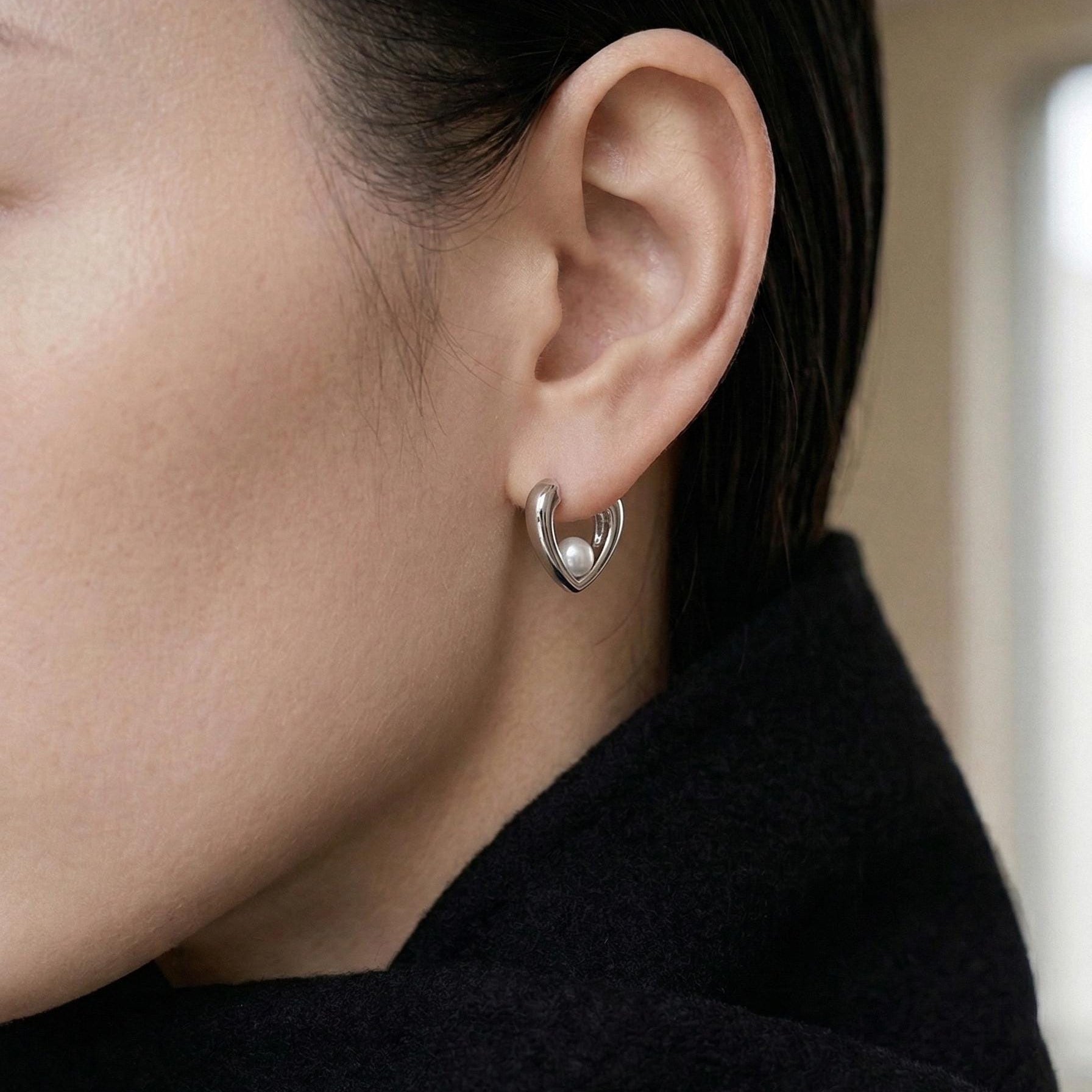 A close-up of a person wearing a Caellia sterling silver heart huggie hoop earring with a pearl.