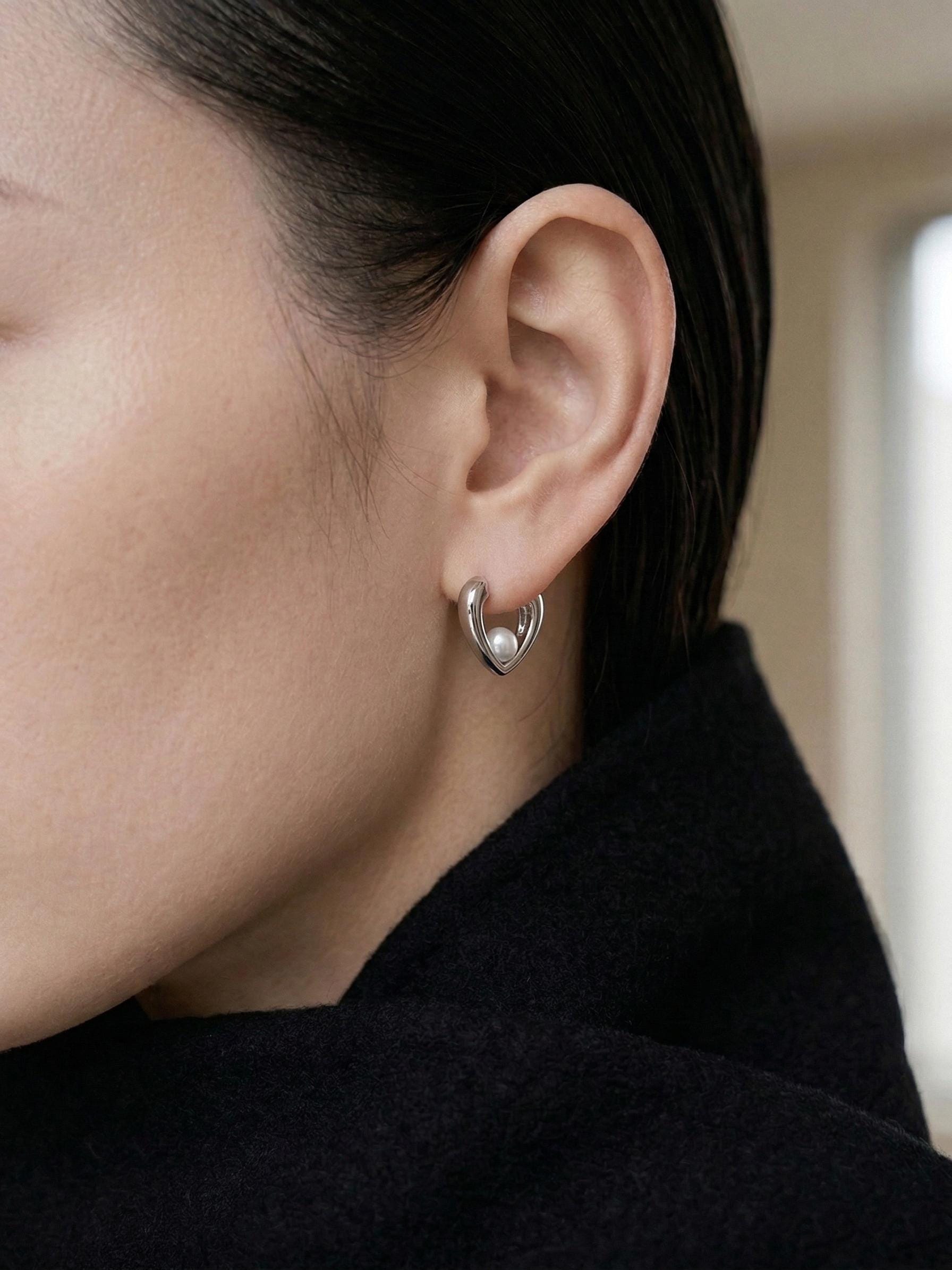 A close-up of a person wearing a Caellia sterling silver heart huggie hoop earring with a pearl.
