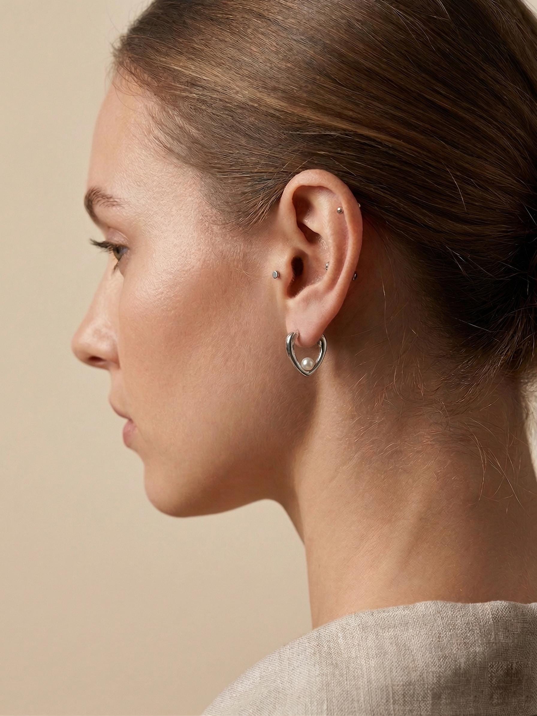 A model wearing Caellia sterling silver heart-shaped huggie hoop earrings with freshwater pearls.