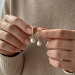 Hands holding a pair of gold half hoop earrings with round white freshwater pearls