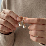 Hands holding a pair of gold half hoop earrings with round white freshwater pearls