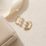 Gold triple pearl earrings arranged beside a cream jewelry gift box